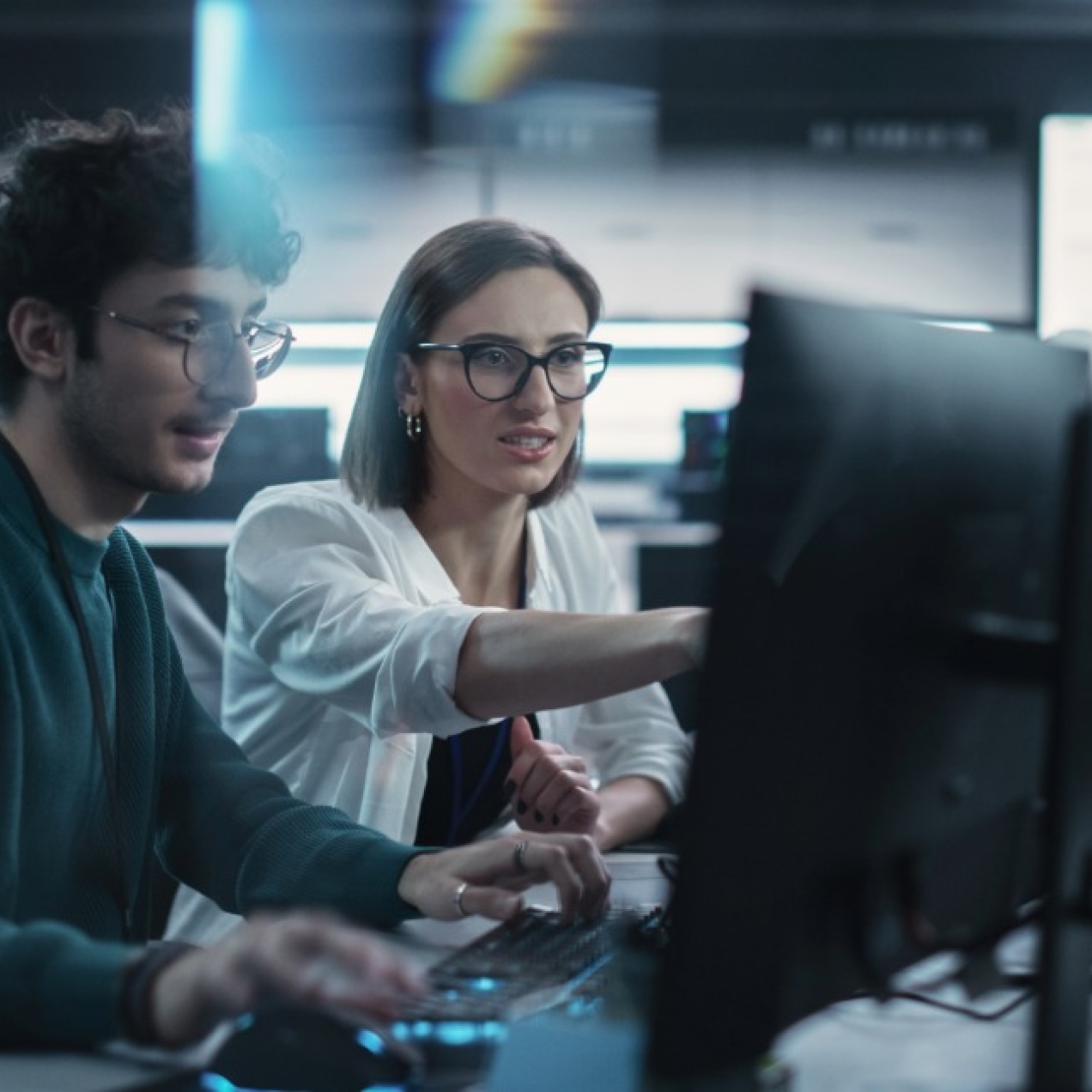 In a modern office a young man and a woman are sat working together at a desk full of computers.